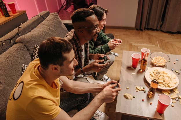 Three young men sit on a couch holding video game controllers; the table in front of them is covered in candy, chips, and popcorn, as well as red SOLO cups and beer bottles.]