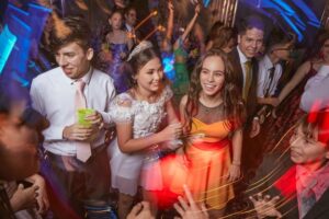 Teens hold beverages and dance at prom. One young woman wears a tiara. The room is illuminated by lights on the dance floor.