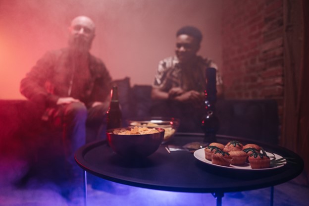 A table in the foreground is covered with snacks, a bottle, and a water pipe used to consume cannabis, tobacco, or other herbal substances. Two men sit on a couch and laugh in the background