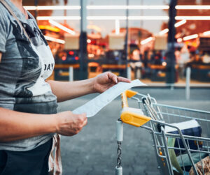 A woman in a t-shirt rests her store receipt on the handle of her shopping cart to examine it in the parking lot