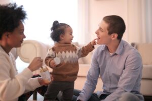 A toddler wearing pants and a sweater stands between her parents who are seated on the floor. The toddler pokes her dad’s lips with one finger while her mother holds a food jar with a spoon.]