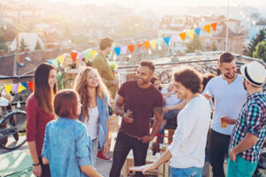 A-group-of-young-adults-gathers-on-a-rooftop-with-string-lights-and-a-colorful-streamer-in-the-background.-Many-of-the-guests-are-holding-beer-bottles-while-more-can-be-seen-on-a-table-nearby