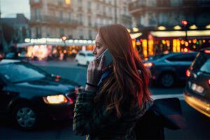 
A-young-woman-talks-on-the-phone-while-standing-on-the-sidewalk-at-night-as-cars-pass-in-the-background