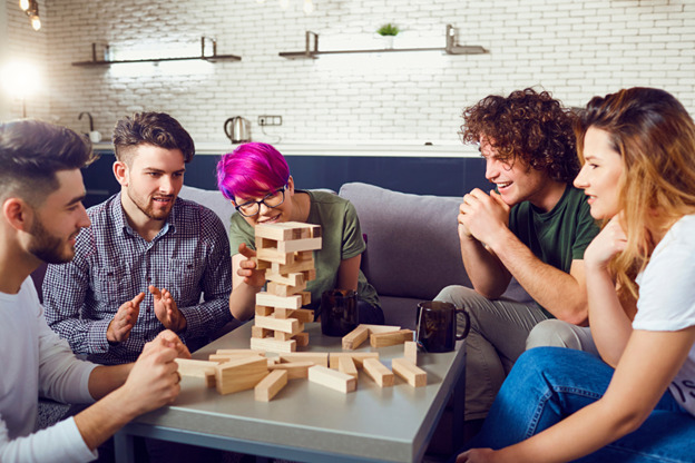 A group of college students gathers around a small table with a Jenga tower. Mugs of tea rest on the table in front of two of them.