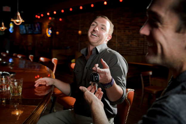 Two men sit at a dimly-lit bar, laughing. One man, with a drink in front of him, is handing his car keys to the other