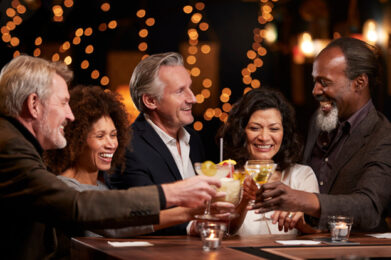 Five middle-aged men and women stand around a bar counter, smiling and laughing. They are clinking their cocktail glasses together