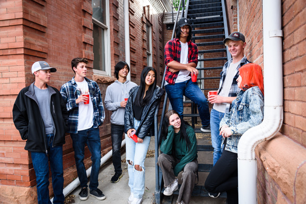 A group of seven teens are gathered outside near a brick building and a metal staircase. They are casually dressed, wearing jeans, hoodies, and jackets. Most are holding red plastic cups and appear to be engaged in conversation. 