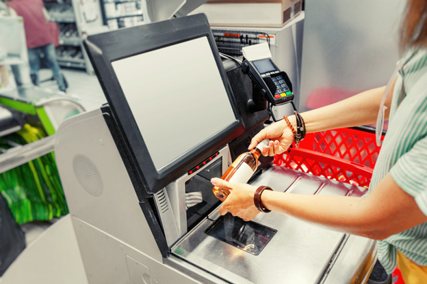 A young woman wearing a white-and-green striped shirt and numerous bracelets scans a wine bottle at a self-checkout register