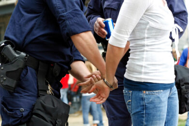 Two police officers in dark uniforms are handcuffing a young person wearing a white top and blue jeans. One officer holds the person's hands behind their back while the other officer holds a beverage can