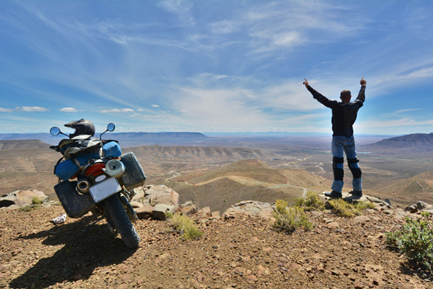 A person is standing triumphantly on a rocky outcrop, with arms raised towards the sky. Next to them is a fully loaded motorcycle