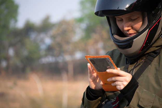 A woman wearing a motorcycle helmet and a green jacket is standing outdoors, looking down at a smartphone encased in an orange cover