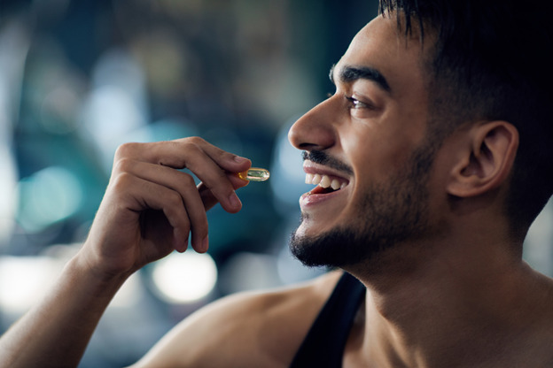 a close-up of a young man smiling, holding a yellow capsule near his mouth, about to take it. He is in a well-lit environment, possibly a gym, as he is wearing a sleeveless athletic shirt. The scene conveys a sense of health or wellness, suggesting the man is consuming a supplement or vitamin as part of a health routine