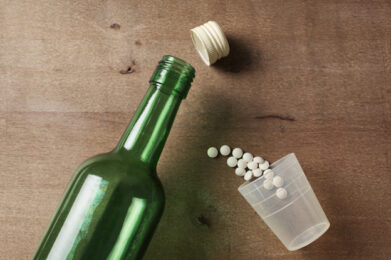 an open green glass bottle lying on its side with the cap detached. Next to the bottle, a clear plastic cup is also tipped over, spilling white round pills onto a wooden surface. The scene suggests a combination of alcohol and medication