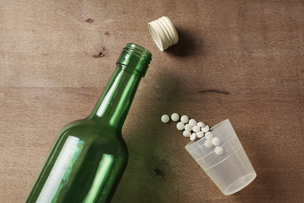 an open green glass bottle lying on its side with the cap detached. Next to the bottle, a clear plastic cup is also tipped over, spilling white round pills onto a wooden surface. The scene suggests a combination of alcohol and medication