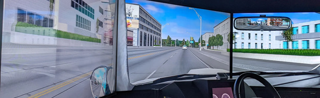 The image shows a driving simulator with a multi-panel screen setup. The view from inside the vehicle's cockpit reveals a city street ahead, with buildings on either side, trees lining the sidewalks, and a few cars in the distance. The rearview mirror in the simulation reflects a clear view of the road behind