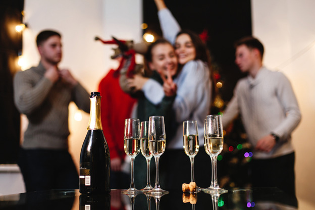 A festive holiday party scene features a bottle of champagne and several filled champagne glasses on a reflective surface in the foreground