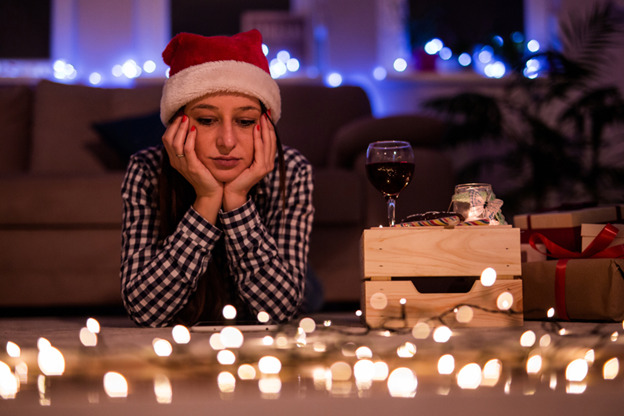 A woman in a checkered shirt and Santa hat sits on the floor with her hands on her cheeks, looking sad. A glass of wine sits on a crate nearby, surrounded by lit candles, gift boxes, and glowing holiday string lights