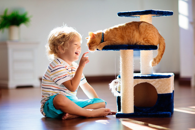 A-young-child-with-curly-blonde-hair-wearing-a-striped-shirt-and-blue-shorts-sits-barefoot-on-a-wooden-floor-smiling-and-reaching-out-to-touch-a-ginger-cats-nose.jpg