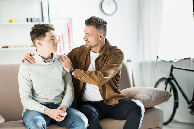 A father and teenage son sit together on a couch in a bright living room, having a warm and supportive conversation. The father, wearing a brown jacket and white t-shirt, has his arm around the son's shoulders and is smiling at him