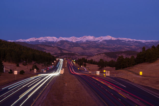 A long-exposure photo of a highway at dusk with light trails from vehicles creating streaks of red and white. The road winds through a mountainous area with snow-capped peaks in the background and forested hills on either side