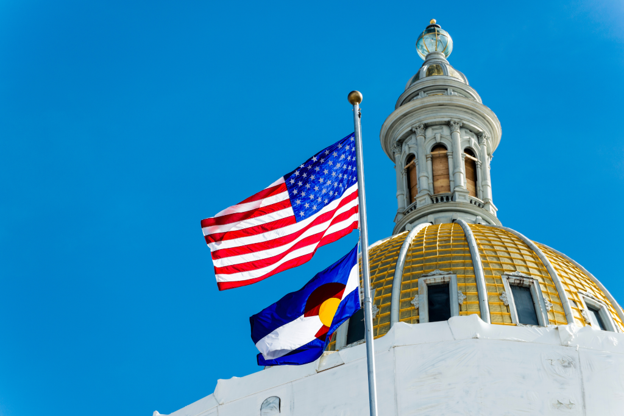 Colorado and U.S. flags flying in front of the gold-domed Colorado State Capitol building against a bright blue sky